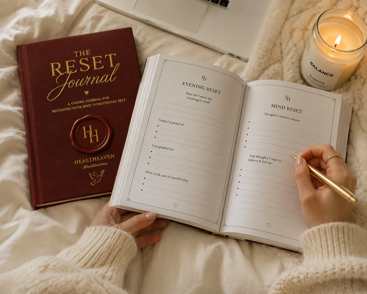 A cozy flat-lay scene showing a maroon hardcover journal titled 'The Reset Journal' next to an open matching planner or notebook with printed pages, held by hands wearing a cream knit sweater. A lit candle in a glass jar labeled 'BALANCE' sits nearby on a soft beige blanket.